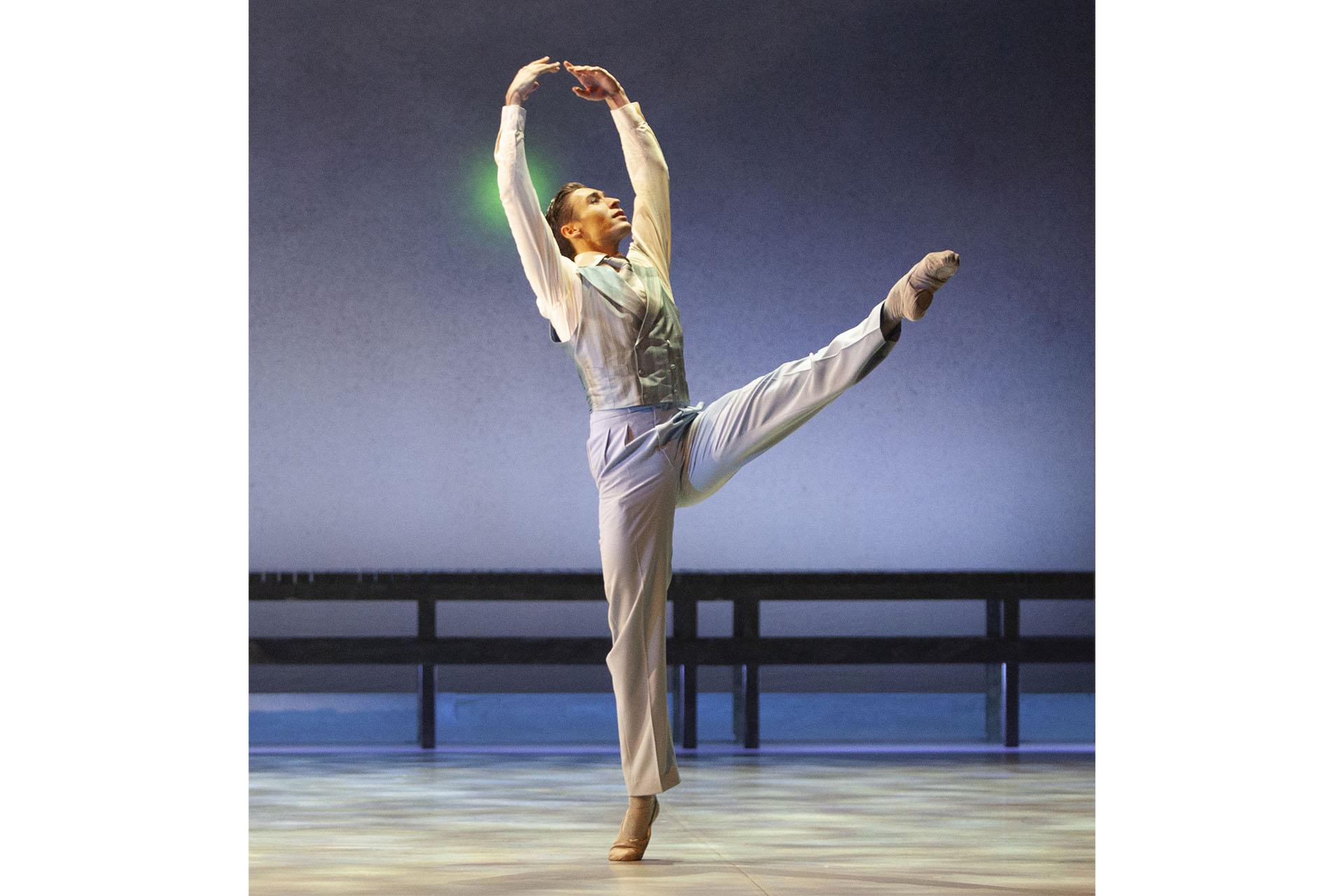 Dressed in a white shirt, pale blue trousers and waistcoat, Jay Gatsby stands on the toes of one foot, lifts his other leg straight in front of him, and fingers of each hand touch at the tips above his head