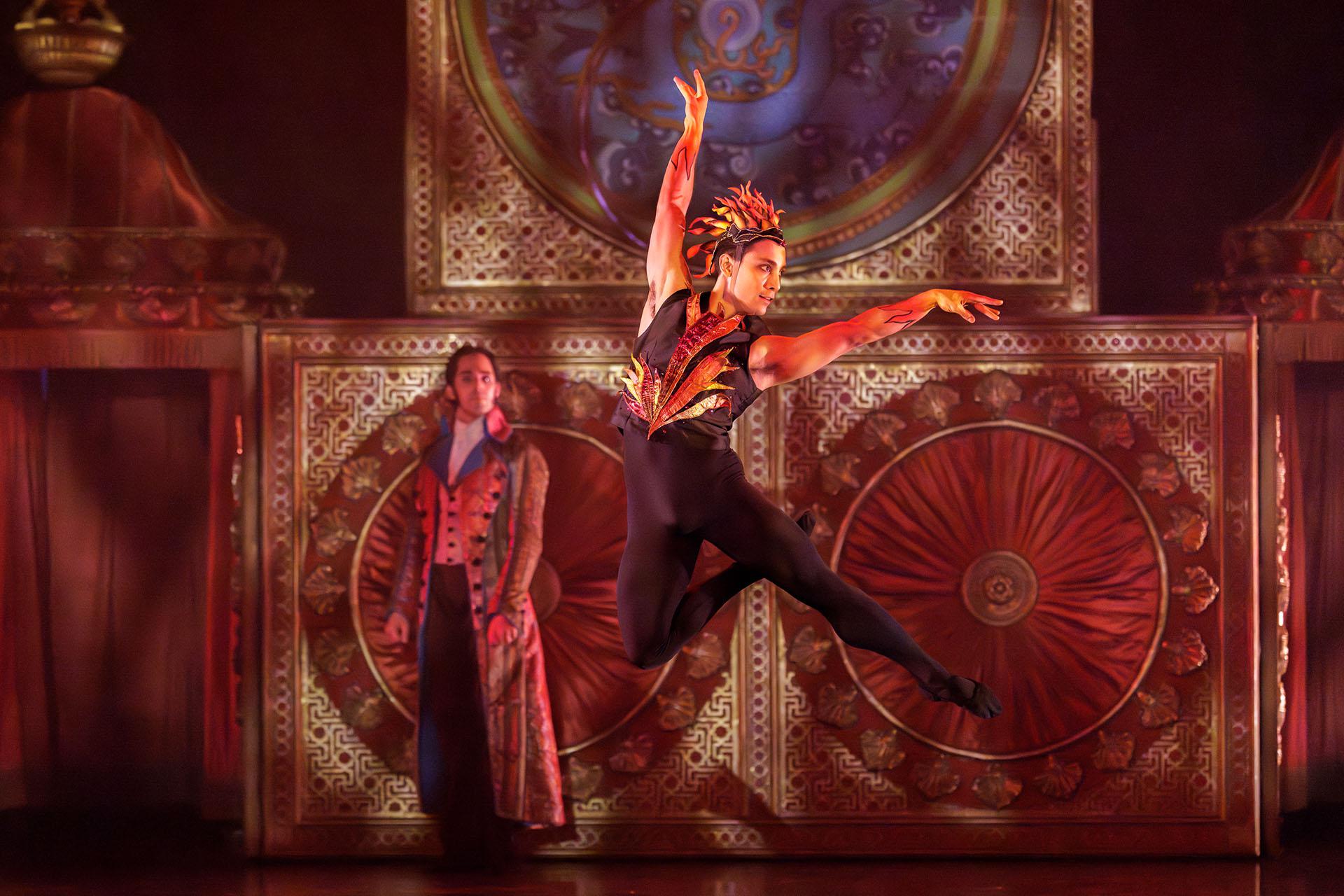 Leaping in the air, man dressed as the element of fire while a stately man looks on