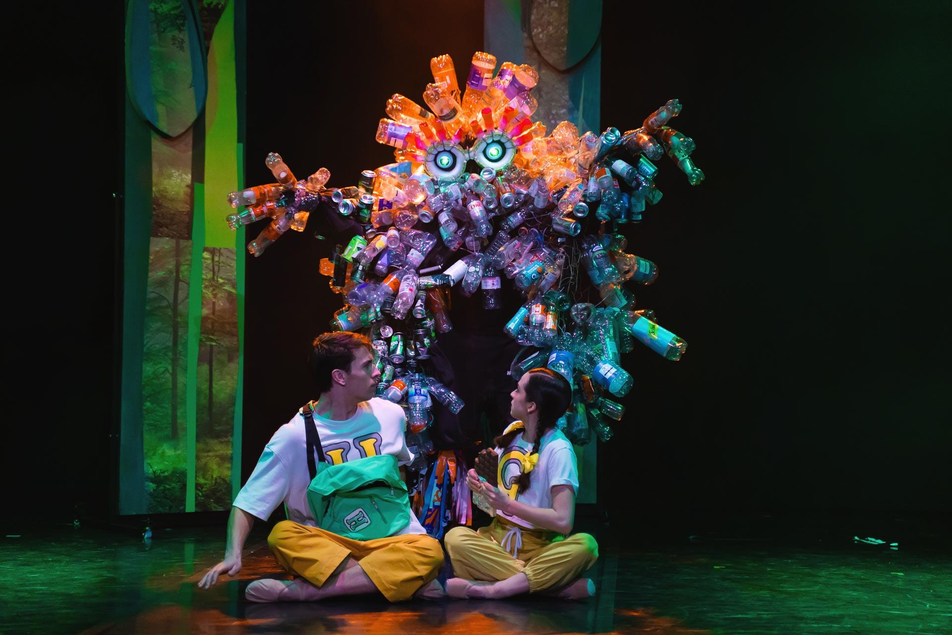 A dancer in a costume covered in plastic bottles sneaking up on two dancers sat at the front of the stage.