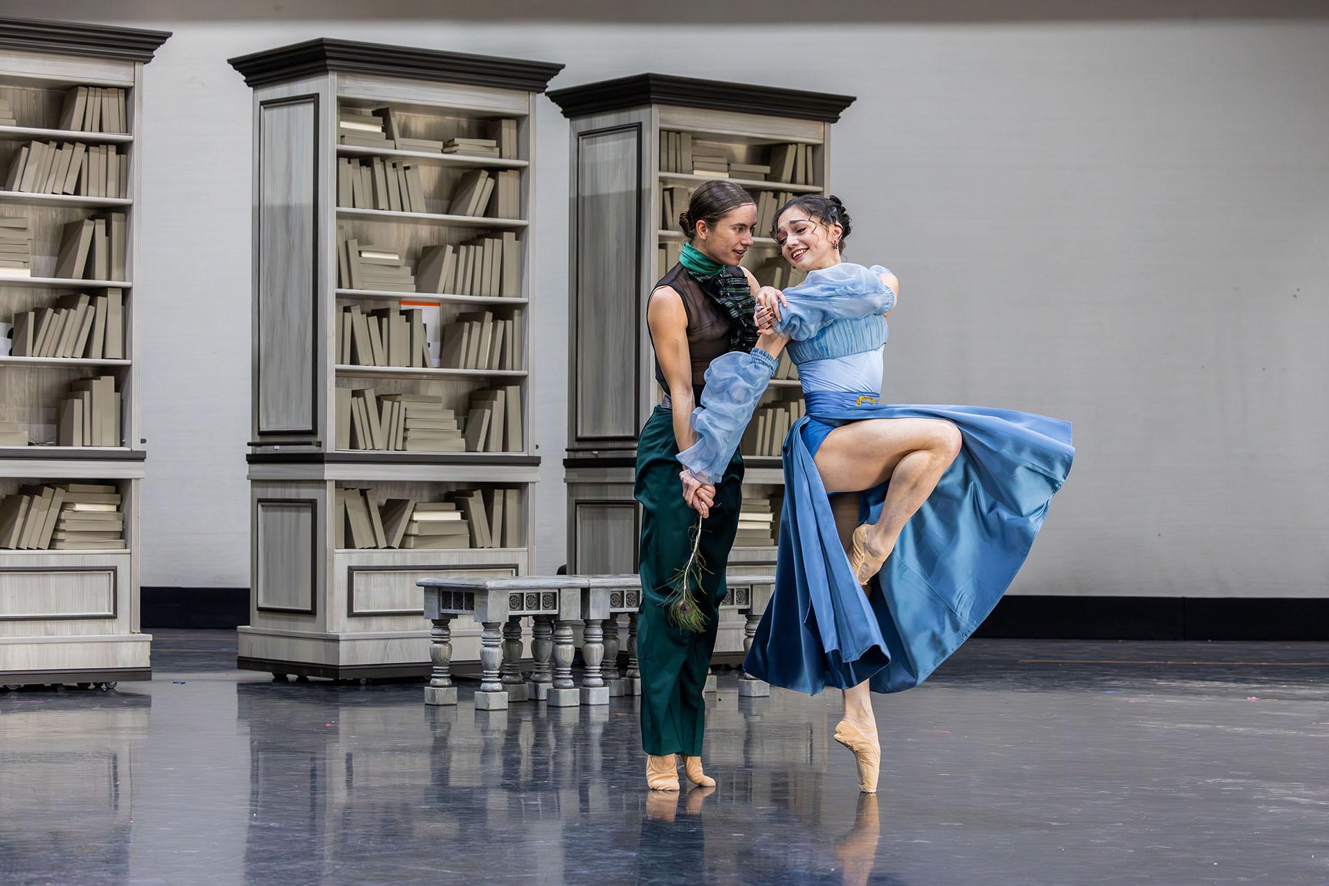 A woman in a blue dress stands en pointe and leans playfully against a woman in dark clothes