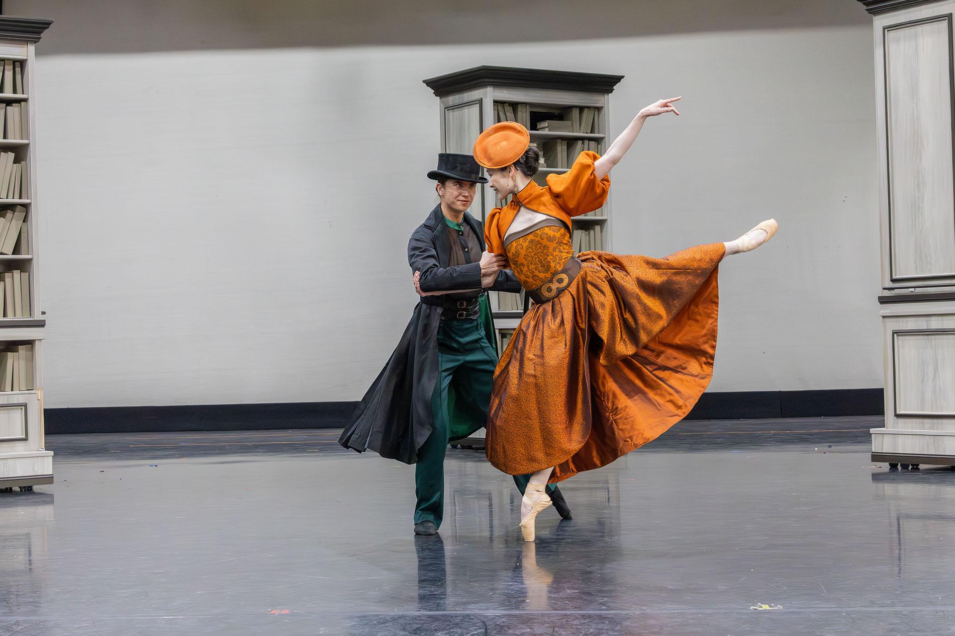 A woman in an orange dress and fascinator dances with Anne Lister who is wearing a hat, long coat and dark green trousers