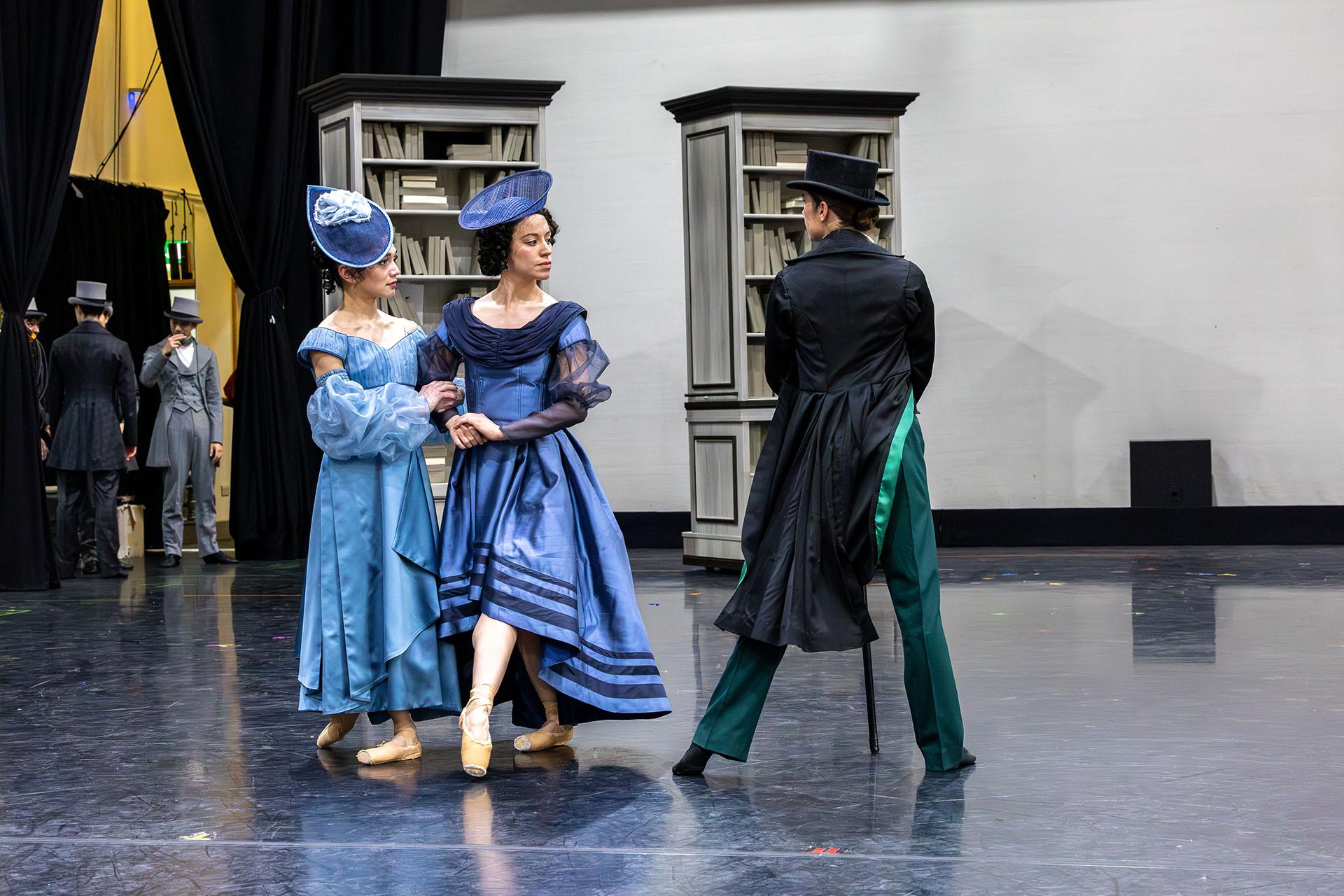 Two women walking arm in arm - one with a pale blue dress, one with a dark blue dress pass Anne Lister wearing a dark long coat and trousers