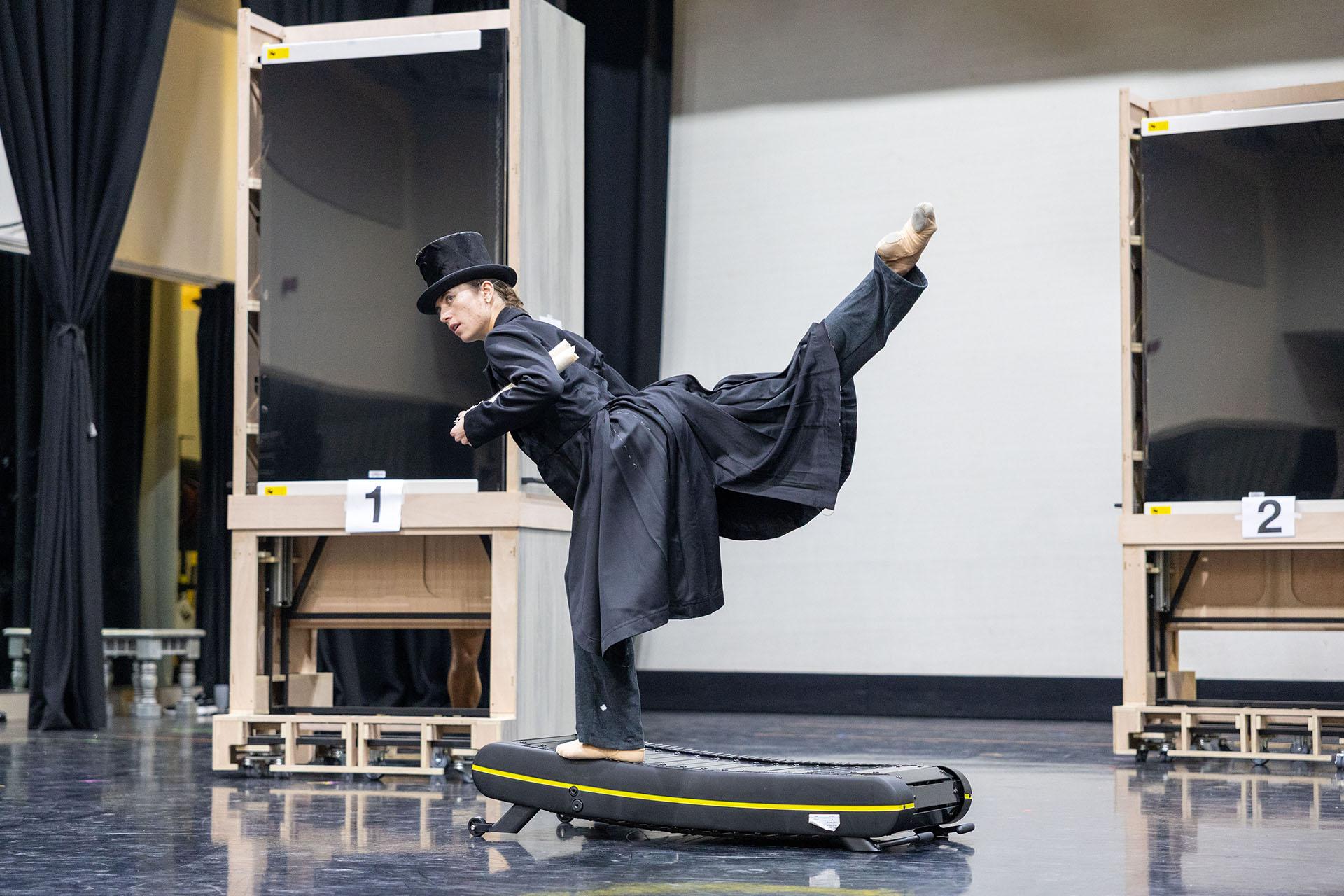 On an automatic treadmill a woman dressed in a long, black coat, top hat and carrying paper under her arms feigns running