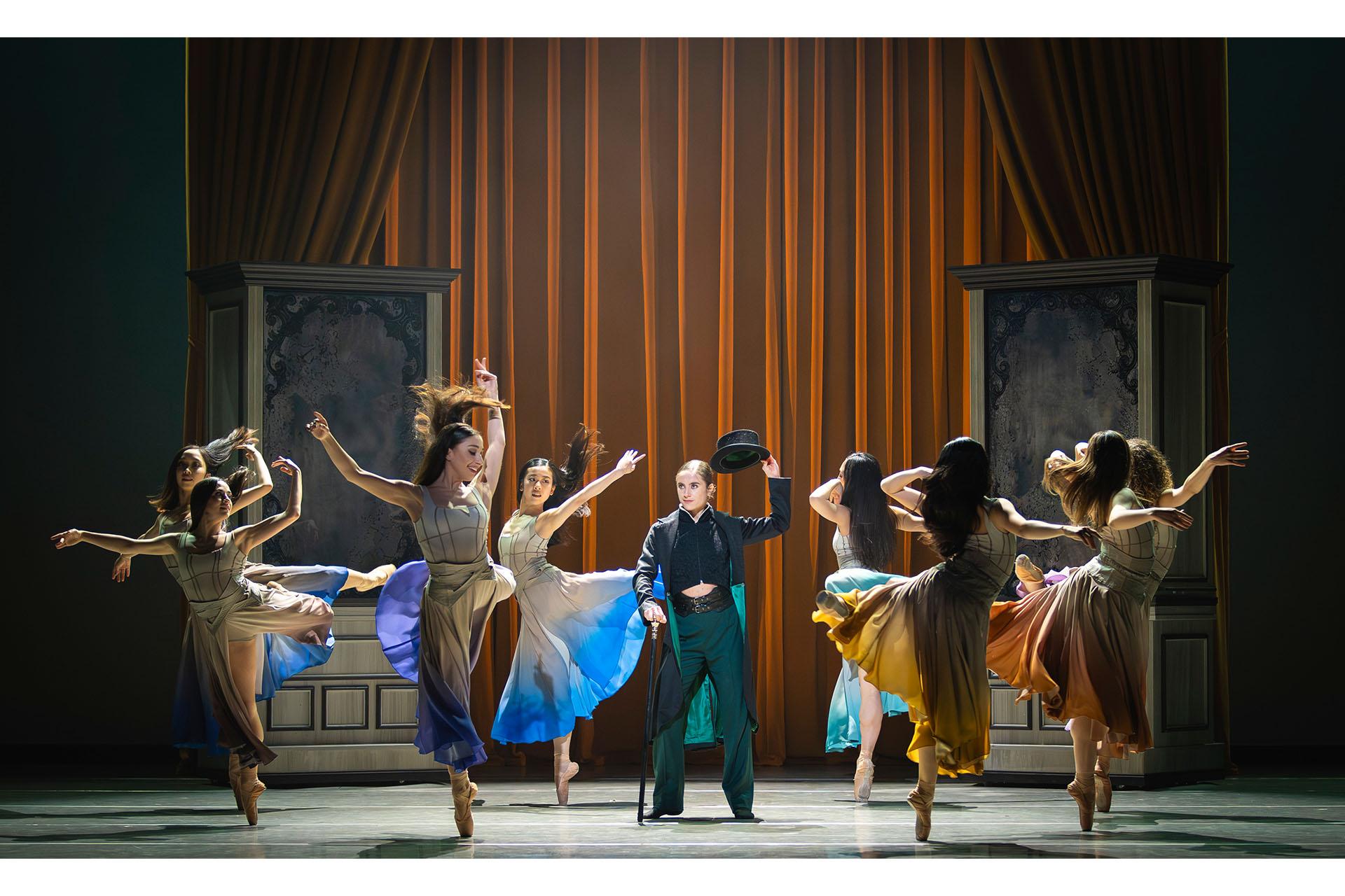 In a dressing room a group of women wearing similar dresses with different accent colours dance around Anne Lister who lifts her hat in respect to them