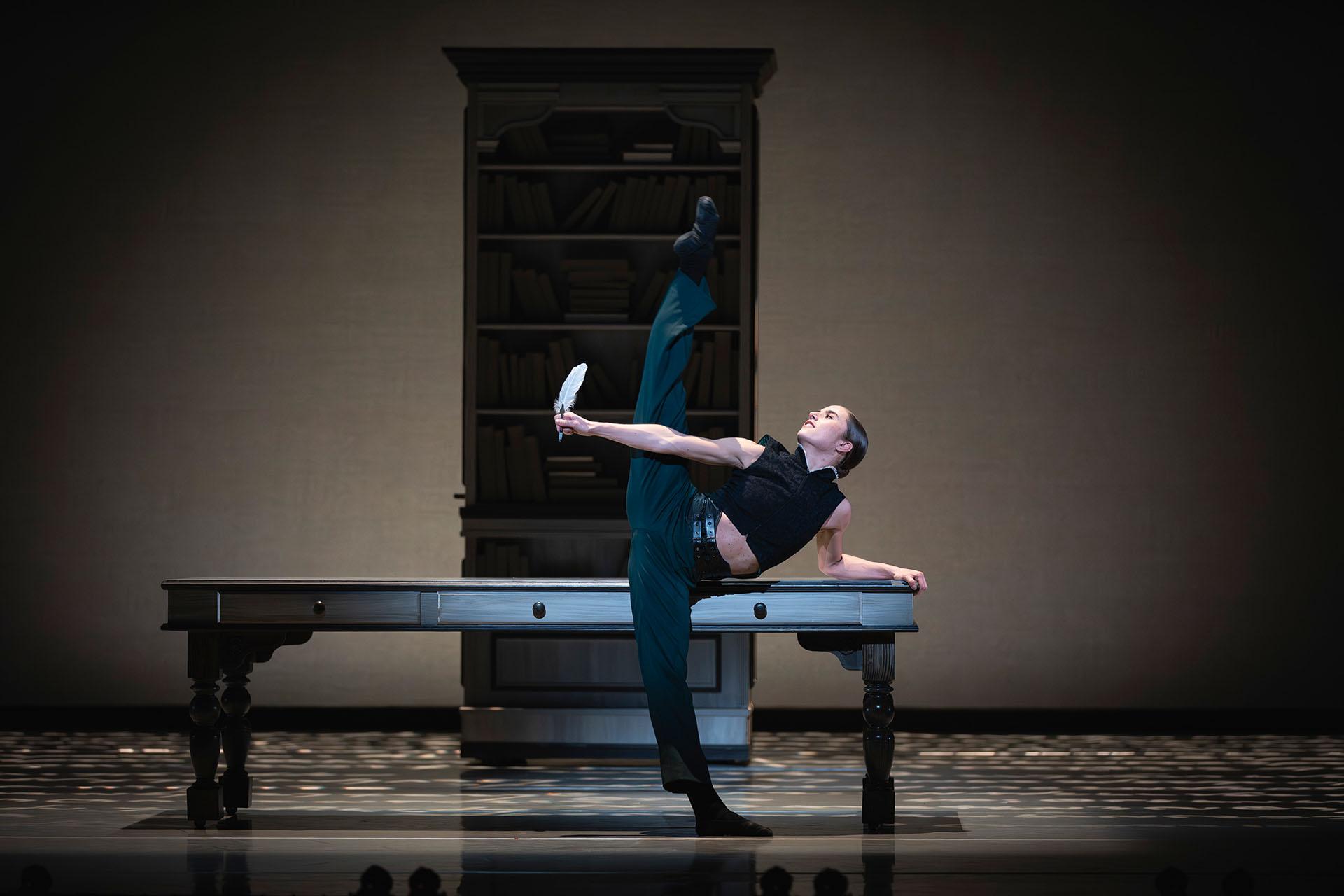 Standing in front of a table, Anne Lister wearing dark green trousers and a back top standing arabesque penché with a quill in one hand