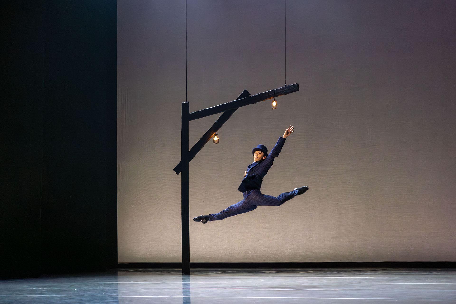 A man in a pin stripe jacket wearing a tall hat leaps across the stage