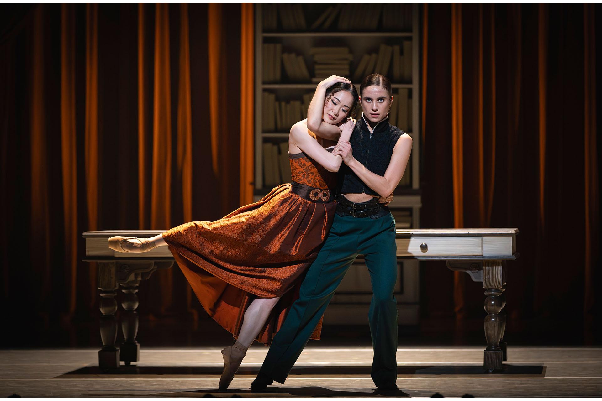 Mariana and Anne Lister stand, holding each other. Mariana in an orange dress, her eye closed as she rests her head on Anne's shoulder, Anne look straight at you