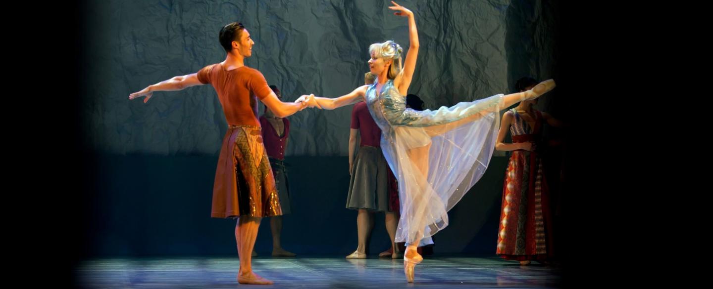 Two dancers on stage, one supports the other as she stands en pointe.