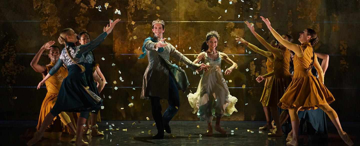 A group of dancers on stage. Two dancers run between two lines of dancers who throw confetti over them in a wedding scene.