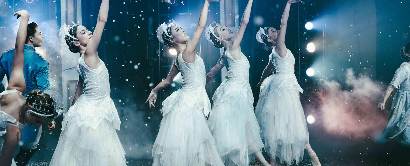 Four female dancers dressed in white each extending a foot down and an arm high