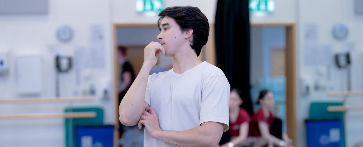 A looks thoughtfully at something in the distance whilst standing in a dance studio.