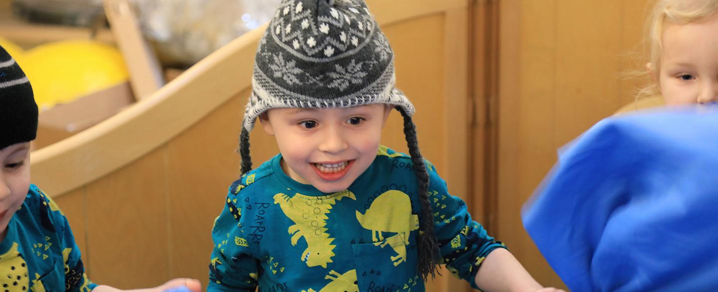 A young child smiling wearing a wooly hat and a green jumper with yellow and black dinosaurs