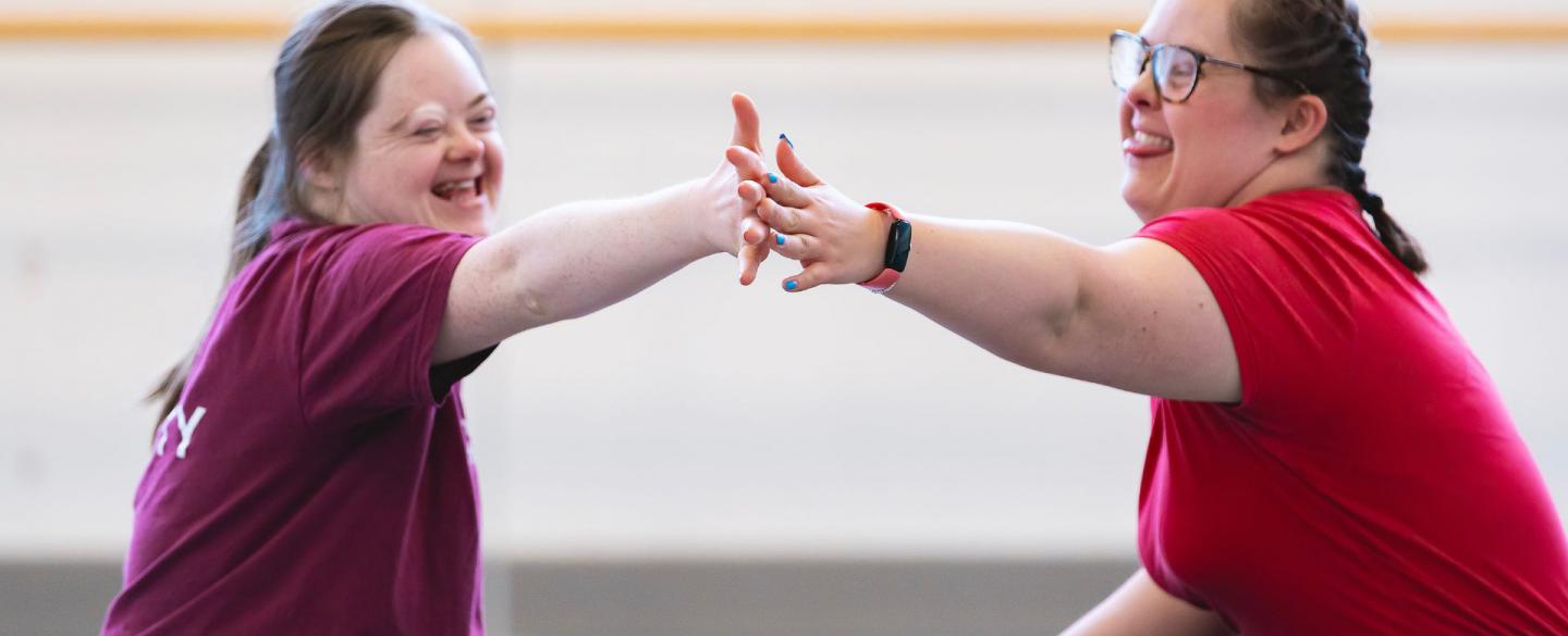 Two dancers in an Ability class sit across from each other on the floor, touching hands and smiling