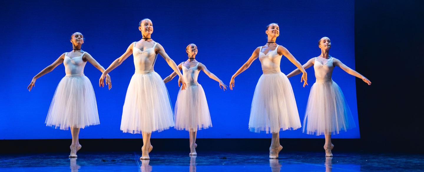 Students in white perform on a stage, standing on their toes with their arms at their side.