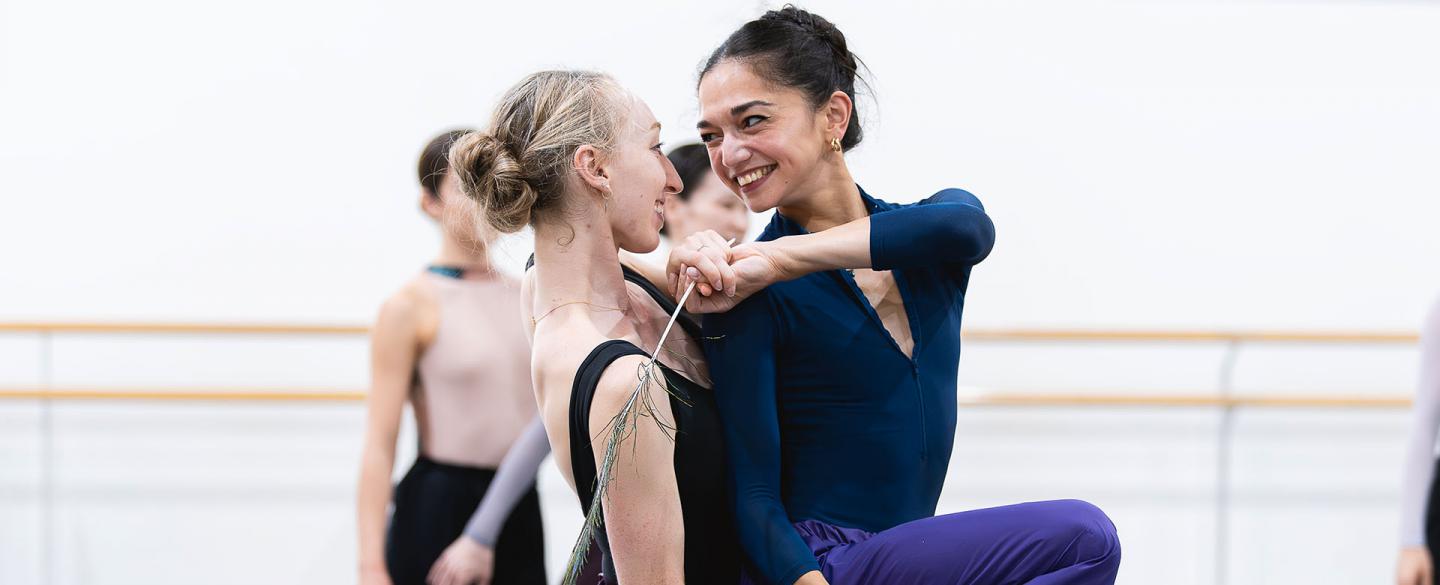 Two dancers looking at one another smiling, the one behind holding the other's leg at the thigh, the one on front reaching around playfully