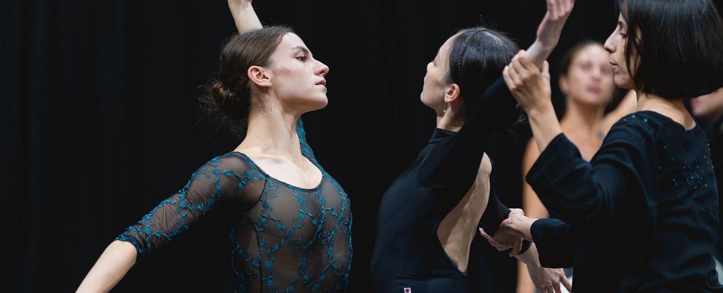 Two dancers face each other, mirroring moves, the choreographer behind one of them guiding her position