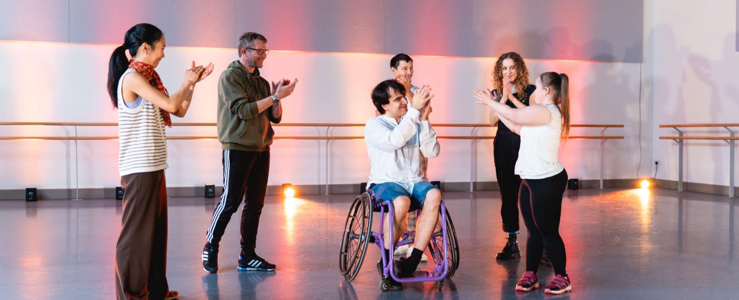 Six people stand infront of warm pink light facing each other, they are applauding.