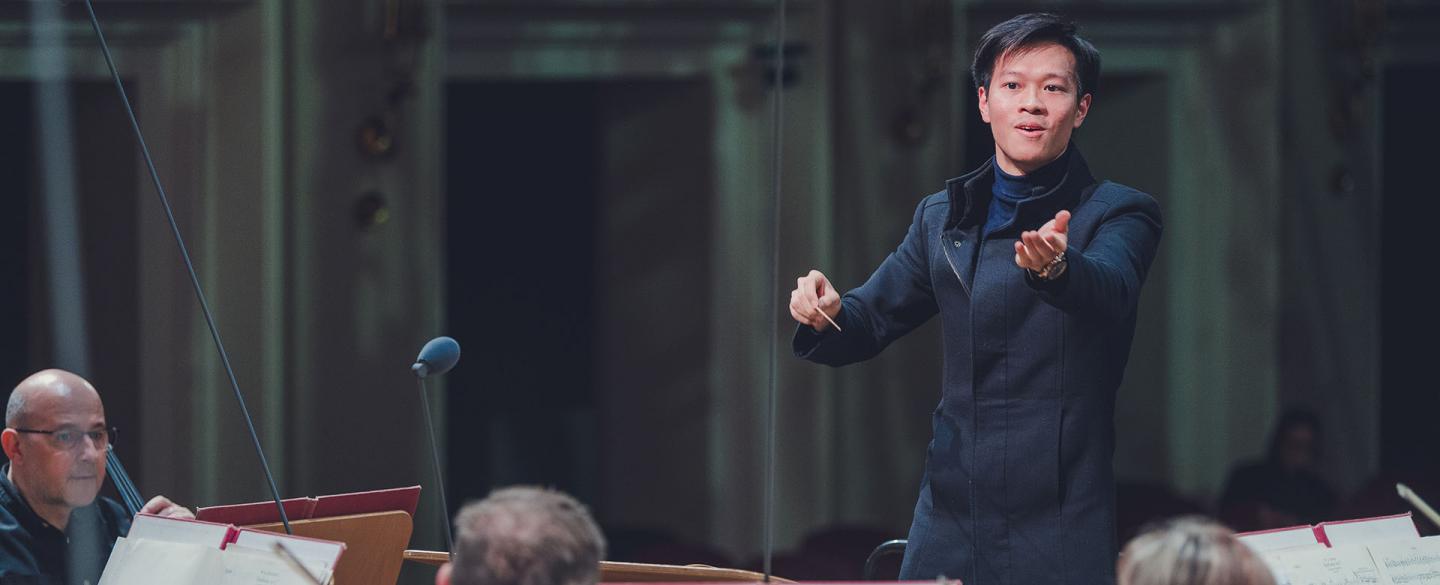 Conductor wearing black, left hand raised to the musicians, right hand holding the baton