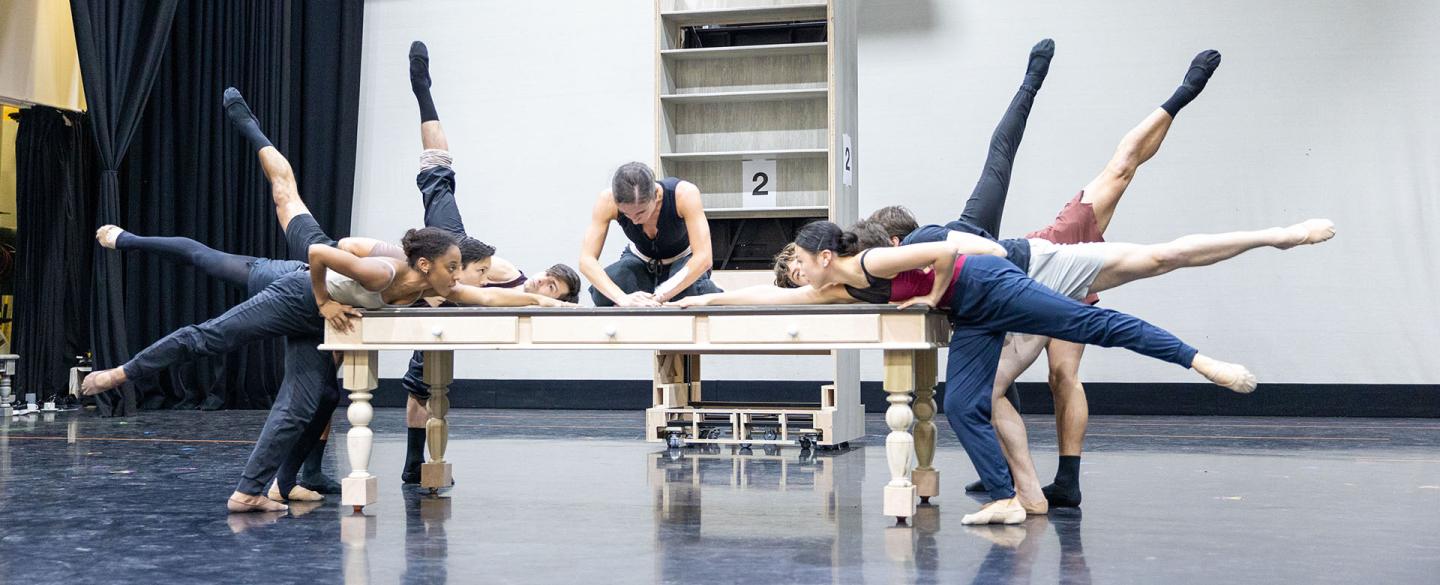 A dancer in black sits on a wooden table, leaning over whilst writing with a feather quill. Multiple dancers surround the table, each with one leg elevated behind them, reaching towards the quill.