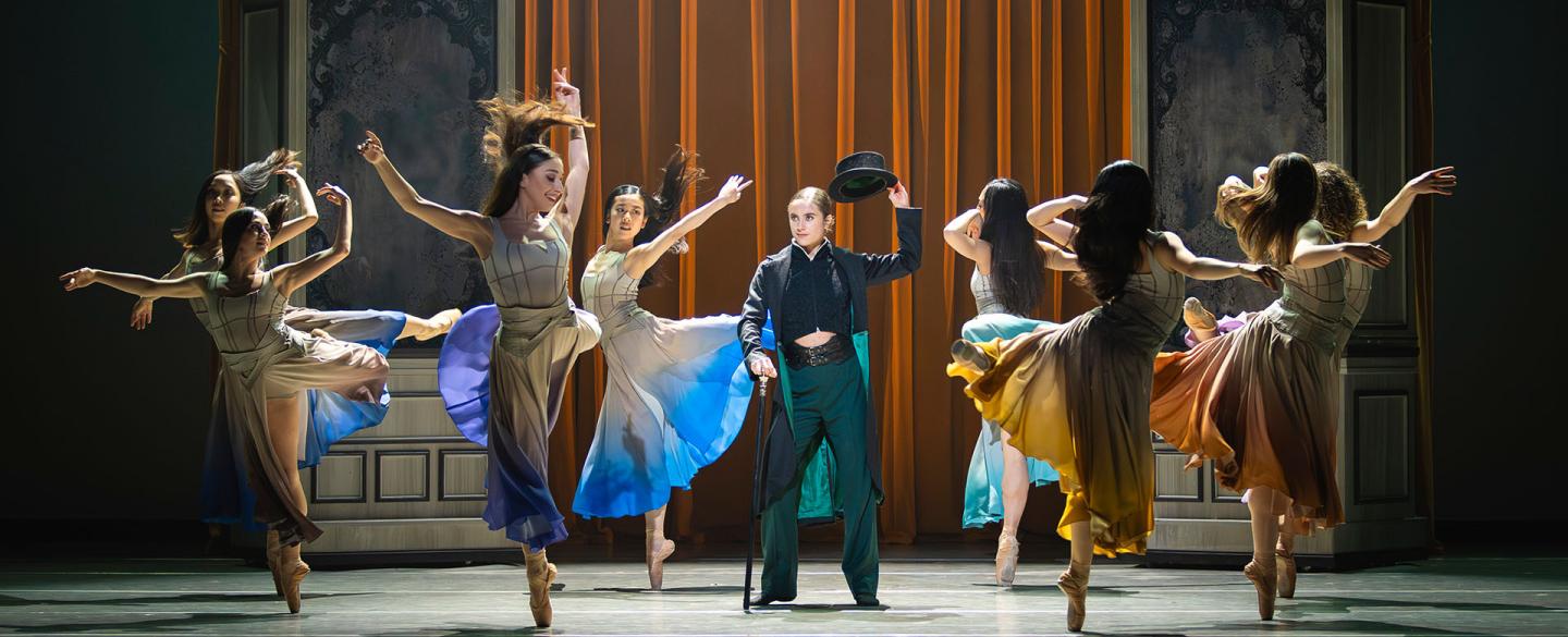 In a dressing room a group of women wearing similar dresses with different accent colours dance around Anne Lister who lifts her hat in respect to them