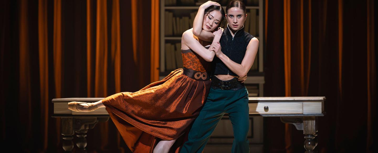 Mariana and Anne Lister stand, holding each other. Mariana in an orange dress, her eye closed as she rests her head on Anne's shoulder, Anne look straight at you