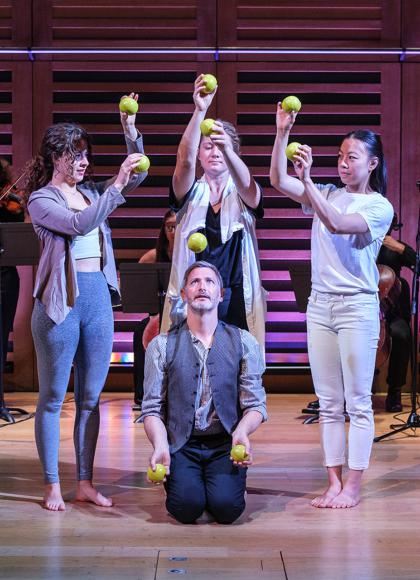 One man juggles while kneeling on the floor while three women stand around him passing and catching balls