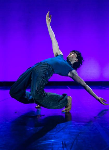male dancer on dark trousers and a tight blue top bends backwards, nearly on the ground but still balanced despite being bent double backwards.