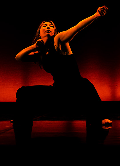 Dancer wearing black trousers and a black vest squat, one arm bent and the other extended as drawing an archery bow.