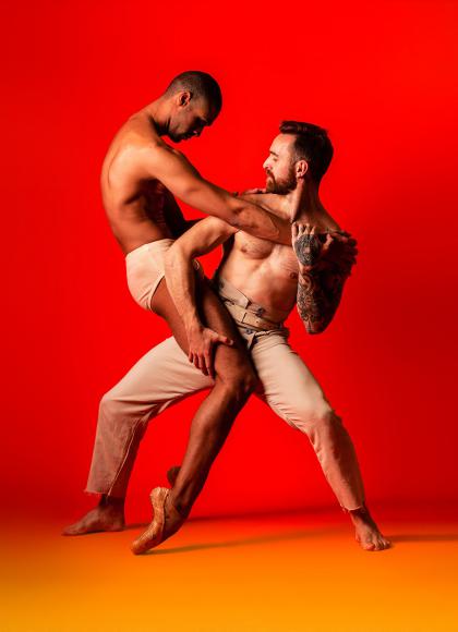 Against a red backdrop, two men dance, embracing each other, simultaneously leaning in an away, wearing pale beige