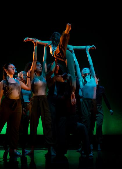 Against a black backdrop, many dancers hold up one with their arms apart, one leg arabesque and the other pointing to the ground