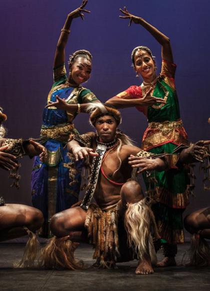 A group of five dancers in traditional costumes strike a pose. Two women are positioned in the centre wearing colourful attire and holding their arms elegantly over their heads. The men, adorned in earthy tones with fur-like accessories, crouch in front. 