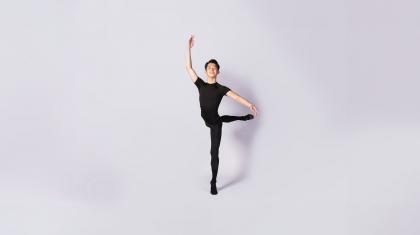 Dancer smiling at the camera, dressed in black, standing on one foot, his other leg raised behind him. His arms are in fourth position, one above his head, the other slightly raised to the side.