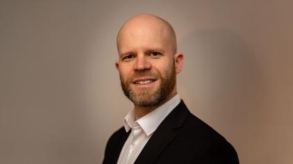 Man with beard smiling at the camera, wearing a black jacket and while shirt, his body in three-quarter profile and head facing us directly