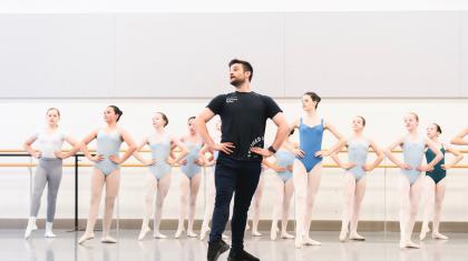 A group of students in a ballet studio stand in a line in front of a barre with their hands on their hips. They're following the lead of teacher Nicola Gervasi who stands in front of them