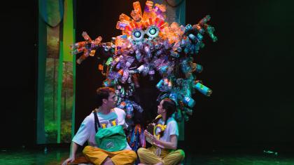 A dancer in a costume covered in plastic bottles sneaking up on two dancers sat at the front of the stage.