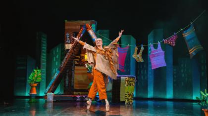 A dancer stood in orange clothes in front of a house with arms outstretched wide