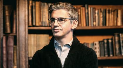 A man stood in front of a wooden bookcase wearing a blue shirt and black jumper. He is wearing glasses and staring off into the distance