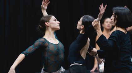 Two dancers face each other, mirroring moves, the choreographer behind one of them guiding her position
