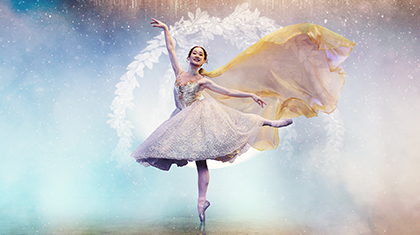 Against a blue and golden fantasy background, a woman stands on one pointed foot, one hand in the air, her other leg balanced elegantly behind her as her dress flows behind her