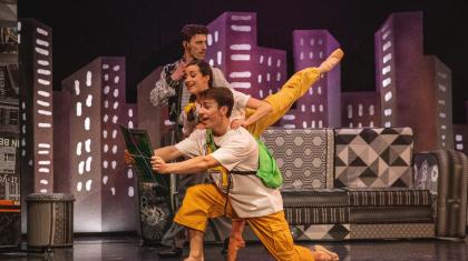 Two dancers look at a map, one over the shoulder of another with leg raised high