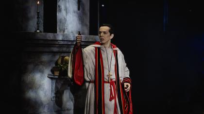 A Friar lifts a dark vial and looks pensive